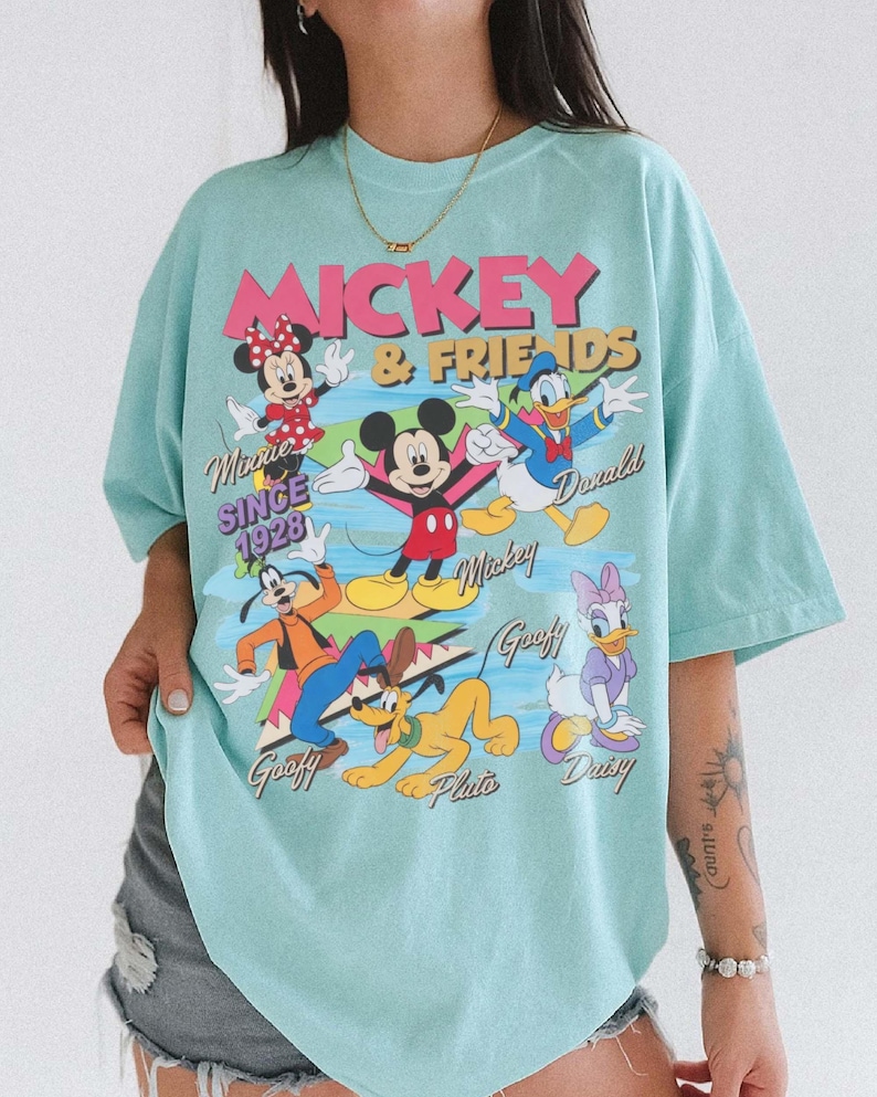 Peut inclure: T-shirt bleu clair avec un motif de Mickey et ses amis. Le graphisme comprend Minnie, Mickey, Donald, Dingo, Pluto et Daisy. Le texte indique "Mickey & Friends" et "Since 1928".