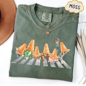 May include: Moss green t-shirt with a graphic of cartoon characters crossing a crosswalk, wearing orange traffic cones. The shirt has a Comfort Colors tag and a gold necklace. The word "MOSS" is in a cloud-shaped bubble.