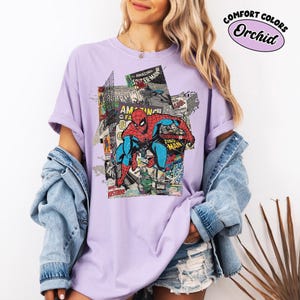 Pode incluir: Uma camiseta lil&aacute;s clara com um design de quadrinhos do Homem-Aranha. O gr&aacute;fico inclui as palavras "Amazing Spider-Man" e "Zing Man". A camisa tem mangas curtas e um ajuste folgado. O texto "Comfort Colors Orchid" est&aacute; no canto superior direito.