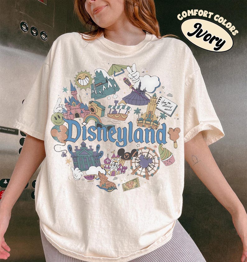 May include: Ivory-colored t-shirt with a Disneyland graphic. The design features various park icons, including a castle, a Ferris wheel, and the Disneyland logo in blue. The shirt is labeled "Comfort Colors".