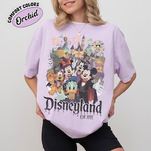 May include: A light purple short-sleeve t-shirt featuring a graphic of Disney characters in Halloween costumes, including Mickey Mouse, Minnie Mouse, Donald Duck, and Pluto. The text reads "Disneyland Est. 1955" with a dripping font effect.