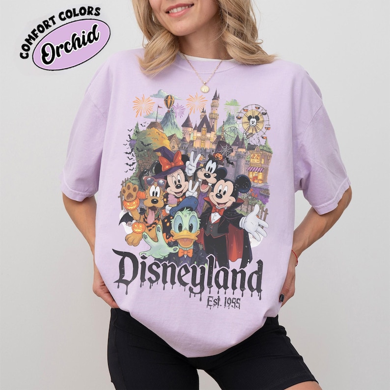 May include: A light purple short-sleeve t-shirt featuring a graphic of Disney characters in Halloween costumes, including Mickey Mouse, Minnie Mouse, Donald Duck, and Pluto. The text reads "Disneyland Est. 1955" with a dripping font effect.