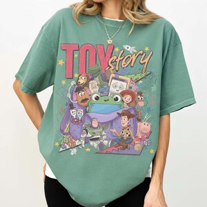 Op de afbeelding: Een saliegroen t-shirt met korte mouwen en een grote grafische print van Toy Story-personages, waaronder Woody, Buzz Lightyear, Jessie, Mr. Potato Head en Rex die uit een paarse rugzak komen, met de tekst Toy Story in dikgedrukte letters.