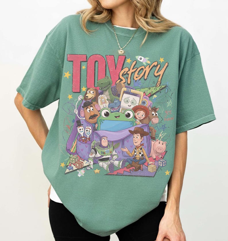 Op de afbeelding: Een saliegroen t-shirt met korte mouwen en een grote grafische print van Toy Story-personages, waaronder Woody, Buzz Lightyear, Jessie, Mr. Potato Head en Rex die uit een paarse rugzak komen, met de tekst Toy Story in dikgedrukte letters.