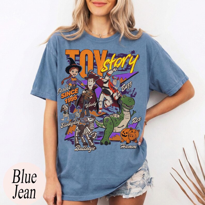 May include: Blue jean colored t-shirt featuring the words "Toy Story" in yellow and purple. The shirt has a graphic of characters from the movie, including Woody, Buzz, and Rex. The text "Jessie Since 1995" is also visible.