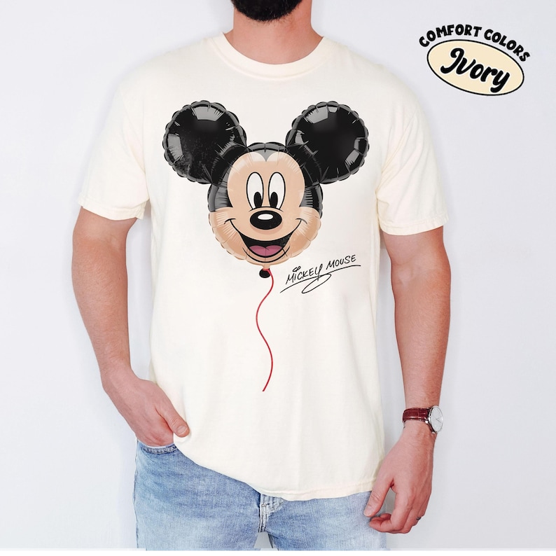 K&ouml;nnte beinhalten: Elfenbeinfarbenes T-Shirt mit einem Mickey Mouse Ballon-Motiv. Der Ballon hat schwarze Ohren und ein l&auml;chelndes Gesicht mit einem roten Faden. Die Worte "Mickey Mouse" sind darunter in Kursivschrift geschrieben.