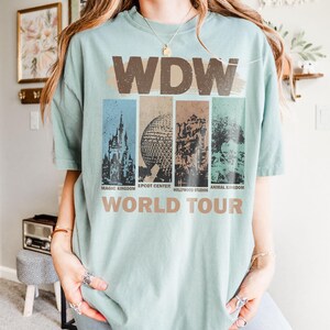 May include: A light blue t-shirt with a vintage-style graphic of the Walt Disney World logo and the text "World Tour". The graphic features four iconic Disney World park icons: Magic Kingdom, Epcot Center, Hollywood Studios, and Animal Kingdom.