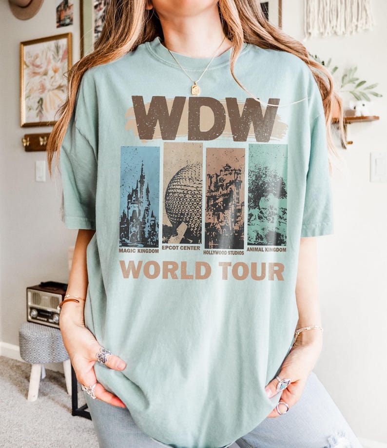 May include: A light blue t-shirt with a vintage-style graphic of the Walt Disney World logo and the text "World Tour". The graphic features four iconic Disney World park icons: Magic Kingdom, Epcot Center, Hollywood Studios, and Animal Kingdom.