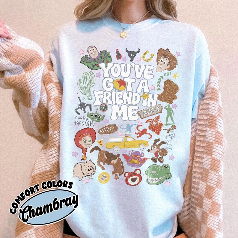 Op de afbeelding: Een lichtblauw T-shirt met ronde hals en een kleurrijke grafische print van diverse Toy Story-personages en iconen. De centrale tekst luidt "YOU'VE GOT A FRIEND IN ME", omringd door illustraties van Woody, Buzz Lightyear, Jessie en Rex.