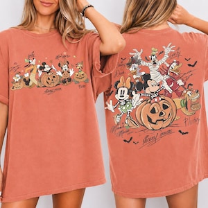 May include: Rust-colored oversized t-shirt featuring a Halloween-themed graphic of Disney characters, including Mickey Mouse, Minnie Mouse, and Goofy, with pumpkins. The back of the shirt also has a similar design. The text "Mickey Mouse" is visible.