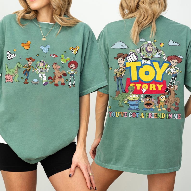 May include: A sage green t-shirt featuring Toy Story characters. The front displays Woody, Buzz Lightyear, Rex, Bullseye, Jessie, and balloons. The back shows the Toy Story logo, characters, and the text, You've Got A Friend In Me.