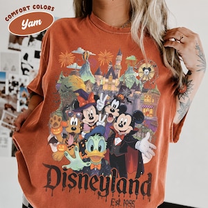 May include: A yam-colored t-shirt featuring a graphic of Disney characters in Halloween costumes, including Mickey, Minnie, Goofy, Donald, and Pluto in front of a castle. The text reads Disneyland Est. 1955 and Comfort Colors Yam.
