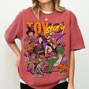 May include: Red-orange t-shirt featuring a graphic of Toy Story characters including Woody, Buzz Lightyear, and Rex. The shirt has the text "Toy Story" and "Jessie Since 1995".