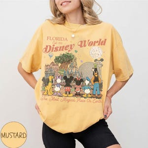 May include: Mustard yellow t-shirt with a retro Disney World graphic. The design shows Disney characters and the text "Florida Disney World Est. 1971" and "The Most Magical Place On Earth".