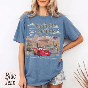 May include: Blue jean t-shirt with a graphic of Radiator Springs from the film Cars. The design includes the text "Radiator Springs California est. 2012" and "Welcome to Radiator Springs" with a red car.