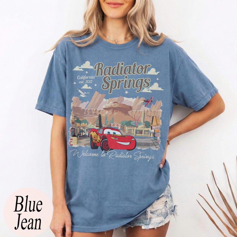 May include: Blue jean t-shirt with a graphic of Radiator Springs from the film Cars. The design includes the text "Radiator Springs California est. 2012" and "Welcome to Radiator Springs" with a red car.
