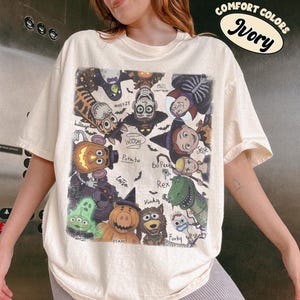 May include: An ivory short-sleeve t-shirt featuring a graphic print of various animated characters dressed in Halloween costumes. The design includes labels for characters like Woody, Buzz Lightyear, Jessie, and Rex, with a Comfort Colors Ivory label.