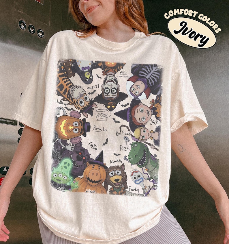 May include: An ivory short-sleeve t-shirt featuring a graphic print of various animated characters dressed in Halloween costumes. The design includes labels for characters like Woody, Buzz Lightyear, Jessie, and Rex, with a Comfort Colors Ivory label.