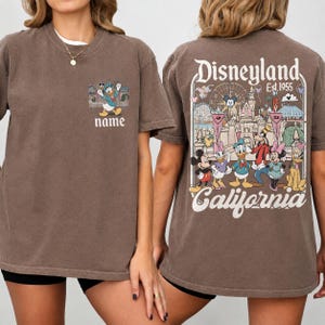 May include: Brown t-shirt featuring a Disneyland design. The front displays Donald Duck with the text "name". The back showcases Disneyland characters and the text "Disneyland Est. 1955 California".