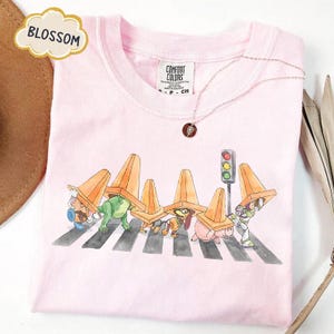 May include: A light pink t-shirt featuring a whimsical illustration of characters crossing a crosswalk, each wearing an orange traffic cone. A necklace with a pendant is visible. The word "BLOSSOM" is in a cloud-shaped graphic.