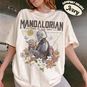 May include: Ivory-colored t-shirt featuring a graphic of The Mandalorian and Grogu surrounded by flowers and butterflies. The text "MANDALORIAN - THIS IS THE WAY" is printed above the image. The shirt also has the text "COMFORT COLORS Ivory" in a circular design.