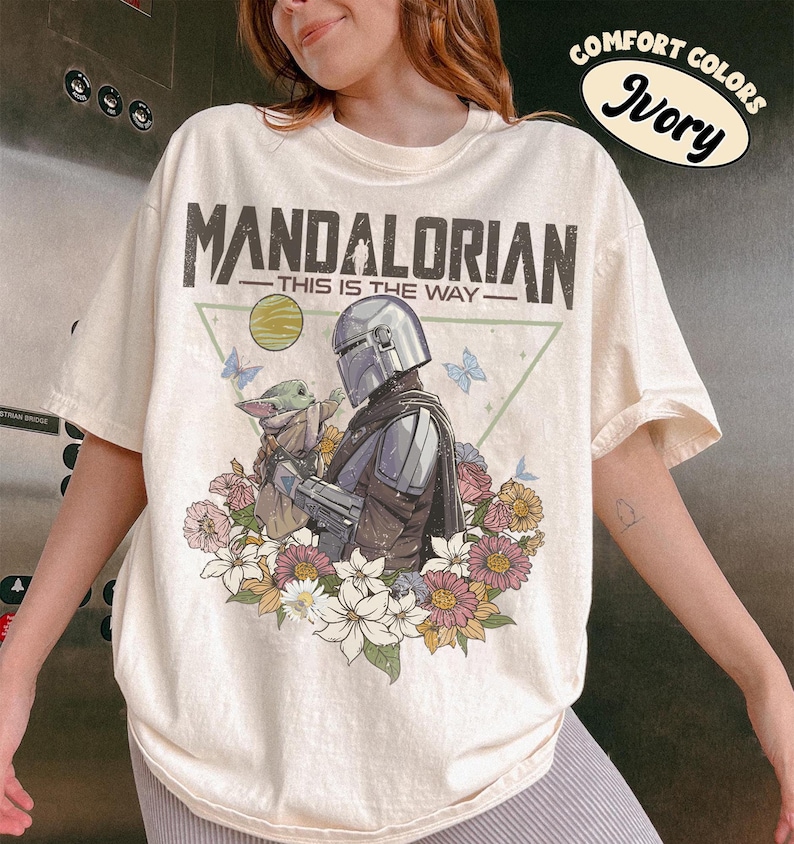 May include: Ivory-colored t-shirt featuring a graphic of The Mandalorian and Grogu surrounded by flowers and butterflies. The text "MANDALORIAN - THIS IS THE WAY" is printed above the image. The shirt also has the text "COMFORT COLORS Ivory" in a circular design.