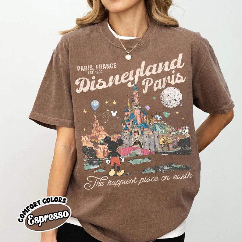May include: Espresso-colored t-shirt featuring a Disneyland Paris graphic. The design includes the text "Paris, France Est. 1992" and an illustration of the castle, Mickey Mouse, and other park elements. The shirt is part of the Comfort Colors collection.