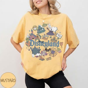 May include: Mustard yellow t-shirt with a Disneyland graphic. The graphic features various Disneyland icons, including a castle, a Ferris wheel, and the Disneyland logo in blue. The shirt is a crew neck and has short sleeves.