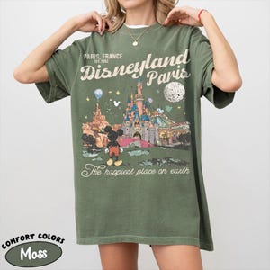 May include: Moss green t-shirt with a graphic of Disneyland Paris, featuring the castle, Mickey Mouse, and the text "The happiest place on earth." The shirt also has the text "Paris, France, EST. 1992."