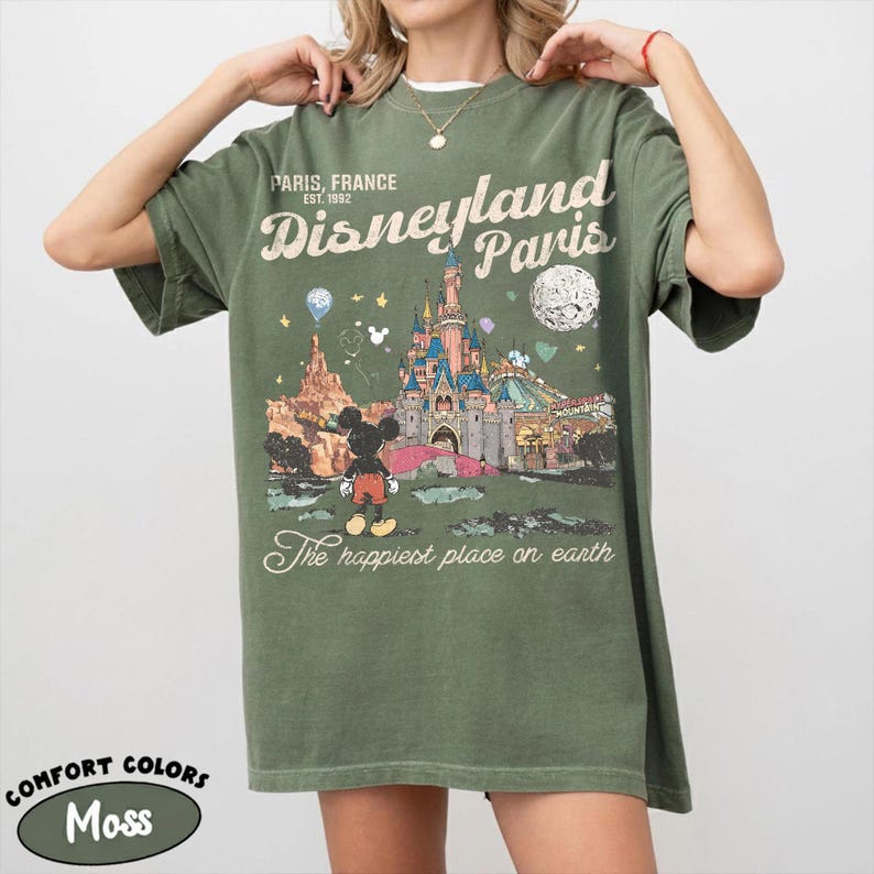 May include: Moss green t-shirt with a graphic of Disneyland Paris, featuring the castle, Mickey Mouse, and the text "The happiest place on earth." The shirt also has the text "Paris, France, EST. 1992."