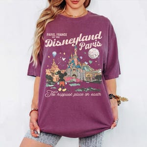 May include: A maroon t-shirt featuring a Disneyland Paris graphic. The design includes the text "Paris, France, Est. 1992", a castle illustration, Mickey Mouse, and the phrase "The happiest place on earth".