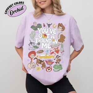 Op de afbeelding: Een lichtpaars t-shirt met korte mouwen en een grafische print met de tekst "YOU'VE GOT A FRIEND IN ME", omringd door diverse kleurrijke illustraties van stripfiguren, waaronder een cowboy, een space ranger, een dinosaurus en een varken.