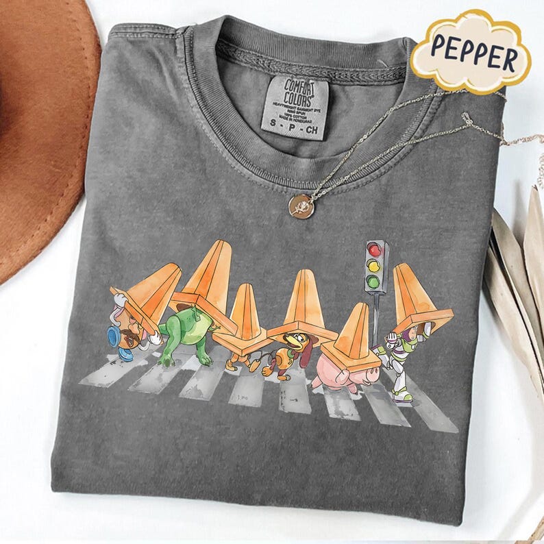 May include: Gray Comfort Colors t-shirt featuring a cartoon illustration of characters crossing a crosswalk, wearing orange traffic cones. A silver necklace and a Pepper tag are also visible.