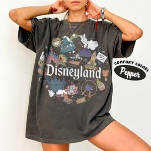 May include: Dark gray t-shirt with a Disneyland graphic. The design features various cartoon illustrations and the word "Disneyland" in white. The shirt also has the words "Comfort Colors" and "Pepper" printed on it.