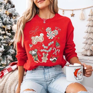 May include: A red sweatshirt with a festive design featuring gingerbread men, candy canes, and the phrase "Tis the Season." The sweatshirt has a crew neck and long sleeves. A white mug with a Santa Claus design is held in the hand.