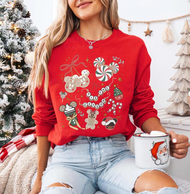 May include: A red sweatshirt with a festive design featuring gingerbread men, candy canes, and the phrase "Tis the Season." The sweatshirt has a crew neck and long sleeves. A white mug with a Santa Claus design is held in the hand.