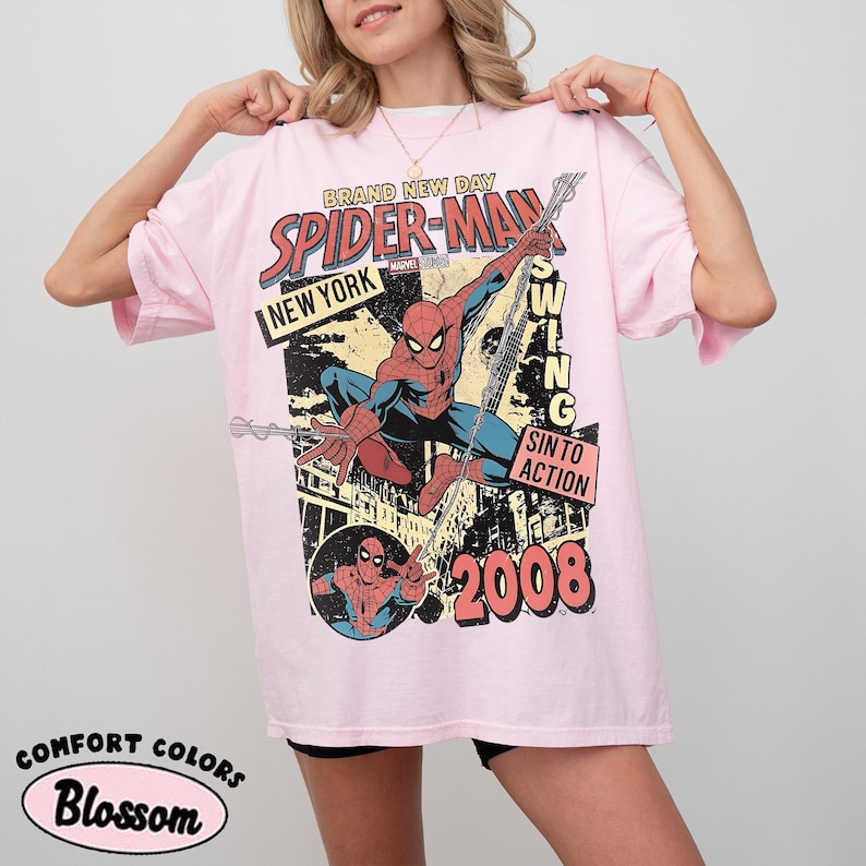 May include: A pale pink t-shirt with a Spider-Man graphic, the text "SPIDER-MAN NEW YORK" and the year "2008". The design also includes "BRAND NEW DAY", "SWING INTO ACTION", and "COMFORT COLORS Blossom".