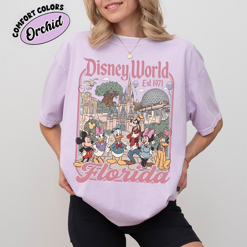 May include: A light purple t-shirt featuring a Disney World graphic with Mickey Mouse, Donald Duck, and other characters. The graphic includes the text "Disney World Est. 1971 Florida" in a pink font. The shirt also has the text "Comfort Colors Orchid".