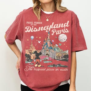 May include: Red t-shirt featuring a Disneyland Paris graphic. The design includes the Disneyland Paris logo, a castle, Mickey Mouse, and the text "The happiest place on earth." The shirt is a vintage style.