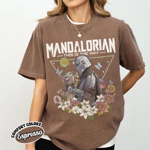 May include: Espresso-colored t-shirt featuring a graphic of The Mandalorian and Grogu surrounded by flowers and butterflies. The text "MANDALORIAN - THIS IS THE WAY" is printed above the image. The shirt is a Comfort Colors brand.