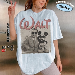 May include: Light blue t-shirt featuring a vintage image of Walt Disney with Mickey Mouse. The word "WALT" is printed in a pink, distressed font above the image. The shirt has a relaxed fit and the words "COMFORT COLORS Chambray" are printed on the top right.