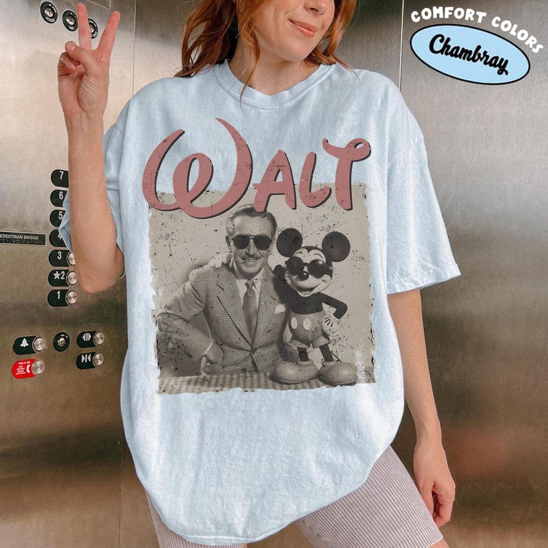 May include: Light blue t-shirt featuring a vintage image of Walt Disney with Mickey Mouse. The word "WALT" is printed in a pink, distressed font above the image. The shirt has a relaxed fit and the words "COMFORT COLORS Chambray" are printed on the top right.