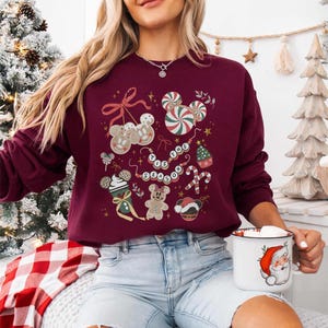 May include: Burgundy sweatshirt with a festive design featuring gingerbread men, candy canes, and the phrase "Tis the Season." The design includes Mickey Mouse-shaped elements and a Santa mug.