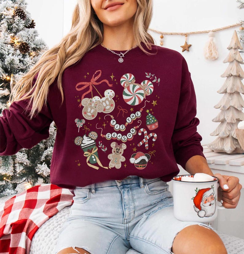 May include: Burgundy sweatshirt with a festive design featuring gingerbread men, candy canes, and the phrase "Tis the Season." The design includes Mickey Mouse-shaped elements and a Santa mug.