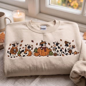 Op de afbeelding: Een beige sweater met ronde hals voorzien van een geborduurd ontwerp van vijf zwart-witte koeien in diverse herfstthema-houdingen, inclusief het dragen van truien en sjaals, zittend bij pompoenen en met koffiebekers tussen bladeren.
