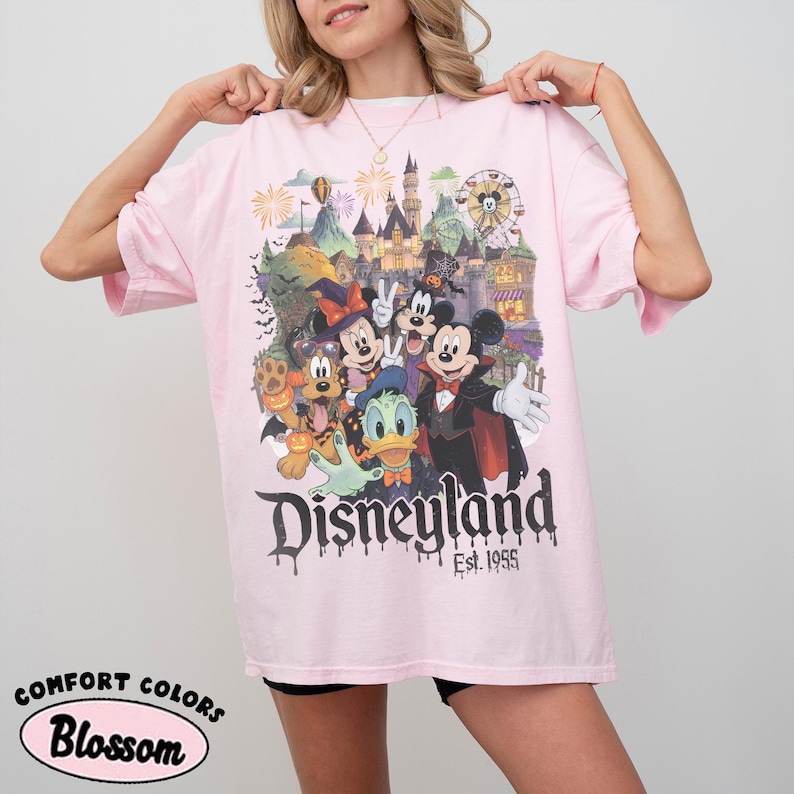 May include: A light pink short-sleeve t-shirt featuring a graphic print of Disney characters in Halloween costumes in front of a castle. The text reads Disneyland Est. 1955. The shirt has a crew neckline and a relaxed fit with a vintage-style design.
