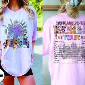Może przedstawiać: R&oacute;żowa koszulka z grafiką World Showcase w Epcot i tekstem "Drink Around the World 19 Tour 82". Grafika zawiera Myszki Miki, Minnie Mouse, Donalda Ducka, Goofy'ego i Pluto. Na plecach koszulki znajduje się lista kraj&oacute;w i miast.
