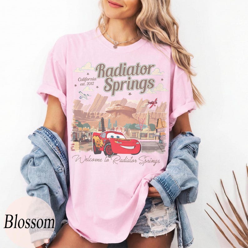 May include: A pale pink t-shirt with a graphic of Radiator Springs from the film Cars. The design includes the text "Radiator Springs" and "Welcome to Radiator Springs" with an image of Lightning McQueen. The shirt also has the text "California est. 2012".