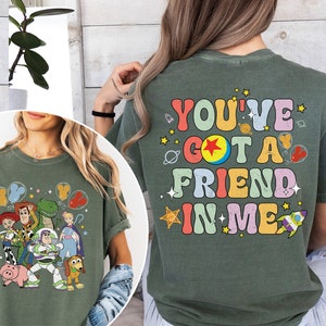 May include: Olive green t-shirt featuring a colorful graphic of Toy Story characters, including Woody, Buzz Lightyear, and Rex. The back of the shirt displays the text "YOU'VE GOT A FRIEND IN ME" in a retro font with space-themed illustrations.