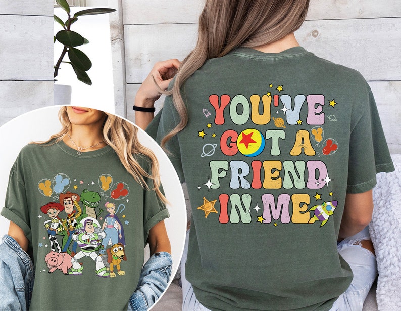 May include: Olive green t-shirt featuring a colorful graphic of Toy Story characters, including Woody, Buzz Lightyear, and Rex. The back of the shirt displays the text "YOU'VE GOT A FRIEND IN ME" in a retro font with space-themed illustrations.